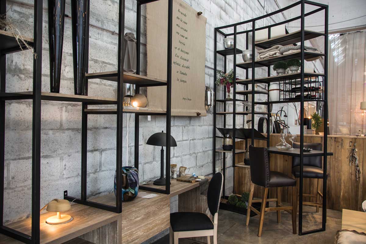 BIBLIOTECA Y BODEGA: dos elementos de usos muy flexibles y visualmente livianos. Biblioteca y bodega semicurva, realizadas en hierro negro y madera, exhiben objetos de deco, de las nuevas líneas que trabaja la tienda: velas, candelabros, vidrios, metales, cuadros, portarretratos, elementos para los toilettes y vajilla para poner espléndidas mesas